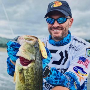 Professional bass fisherman Randy Howell holds up a fish with a yellow Livingston lure in its mouth.