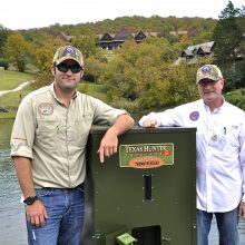 Kyle Moon and Phillip Moon standing next to a Texas Hunter fish feeder beside a lake at Big Cedar Lodge.